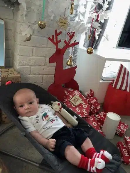 Sebby sits in his bouncinet, wearing a white tshirt, and dark blue shorts. Sebby is awake and staring at the camera. He has a Rudolf the Red-Nosed Reindeer rattle lying next to him. Sebby is wearing red and white striped Santa socks. In the background are presents and Christmas decorations.