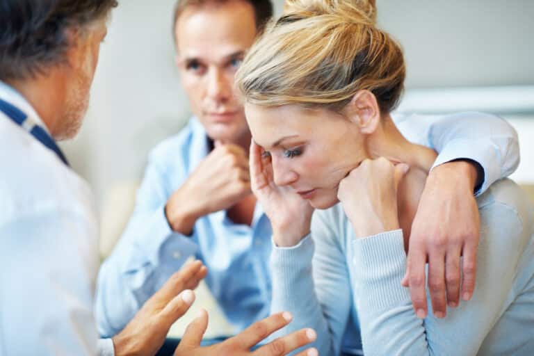 Image is of a couple in a doctor’s office. Both the man and woman look sad.