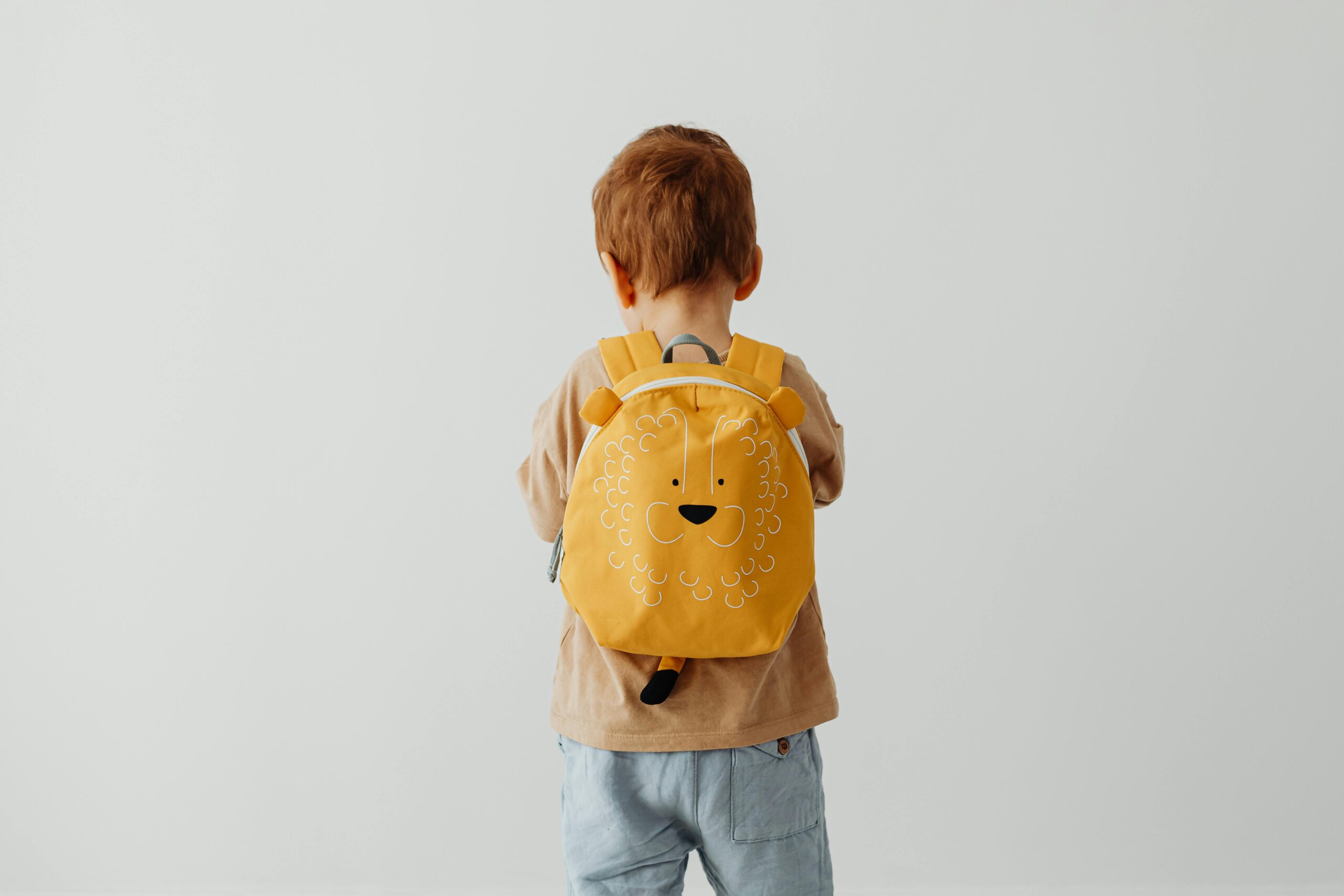 Cute child with red hair wearing a playful lion backpack facing away.