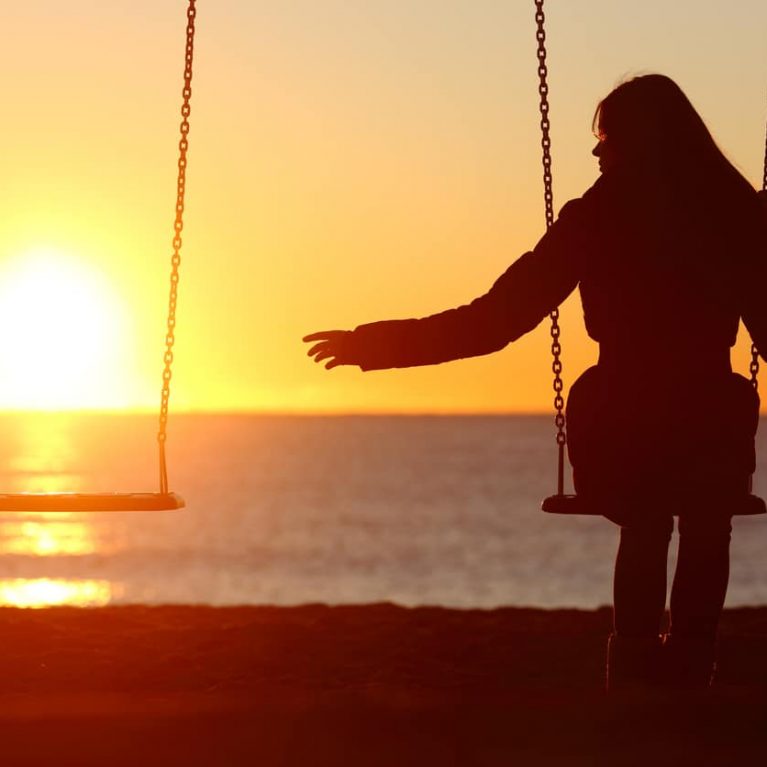 Silhouette of a girl on a swing at sunset, reaching towards an empty swing beside her, for Bereaved Siblings Month
